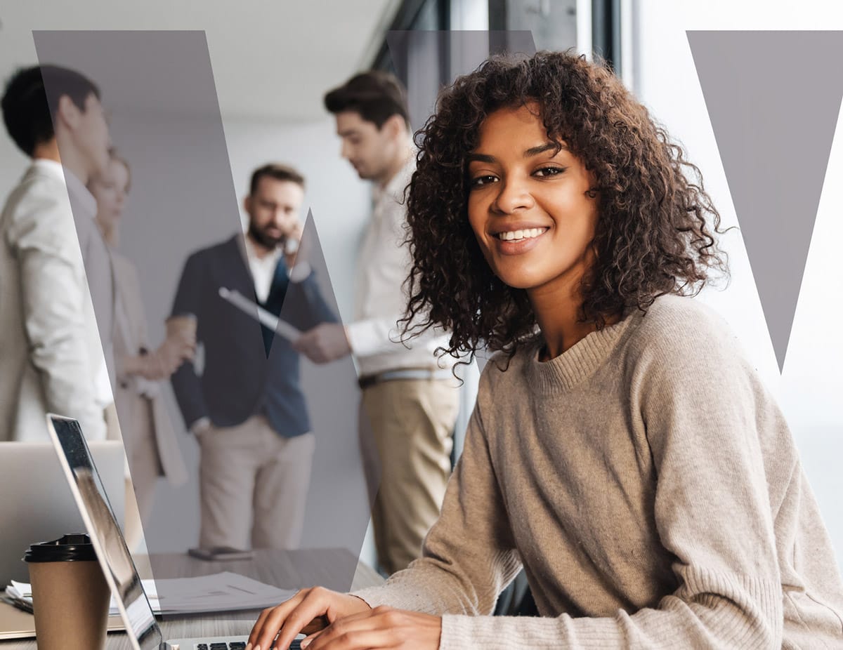 Smiling woman working on a laptop in a modern office, with colleagues collaborating in the background.
