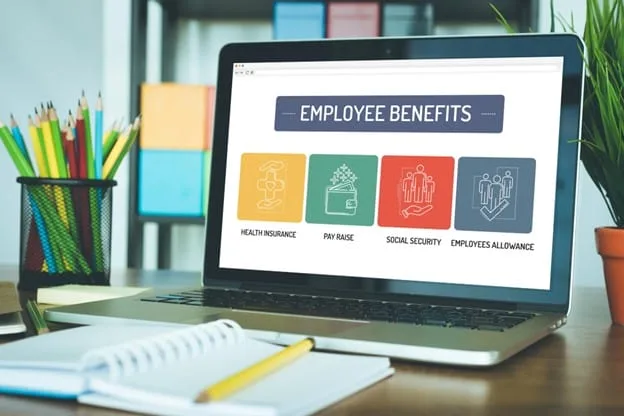 A laptop computer rests on a desk in front of an open spiral-bound notebook and a wooden pencil. The laptop screen says, “EMPLOYEE BENEFITS” across the top and displays graphics labeled “HEALTH INSURANCE,” “PAY RAISE,” “SOCIAL SECURITY,” and “EMPLOYEES ALLOWANCE” below.