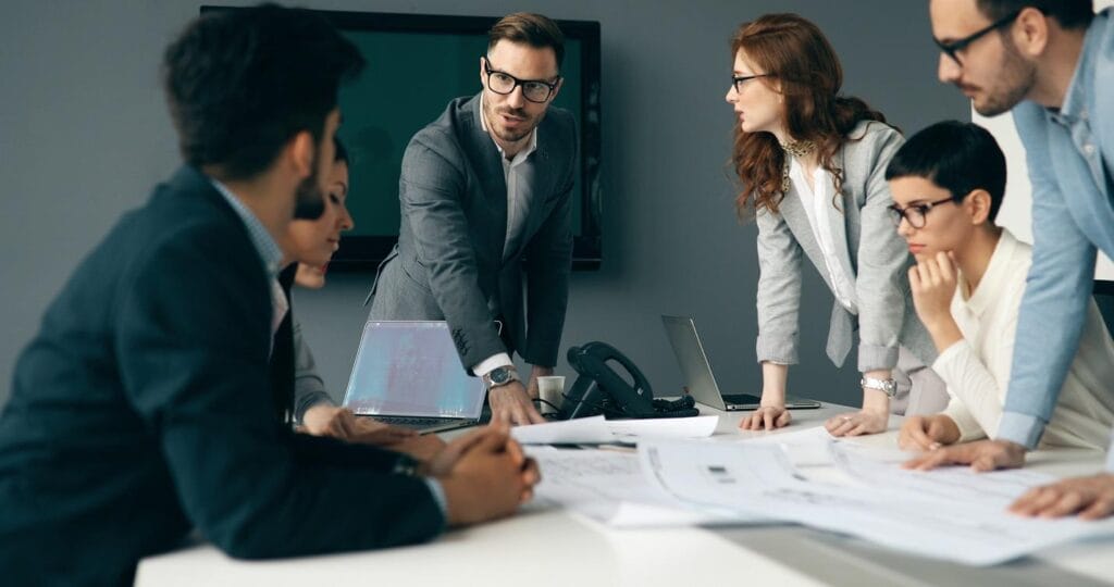 Business team in conference room; mixed-gender business team in suits sitting and standing around a conference table; captive insurance strategy session.