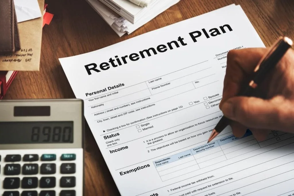 A calculator sits on a desk beside a paper that is titled Retirement Plan and a hand holding a pen hovers over the paper.