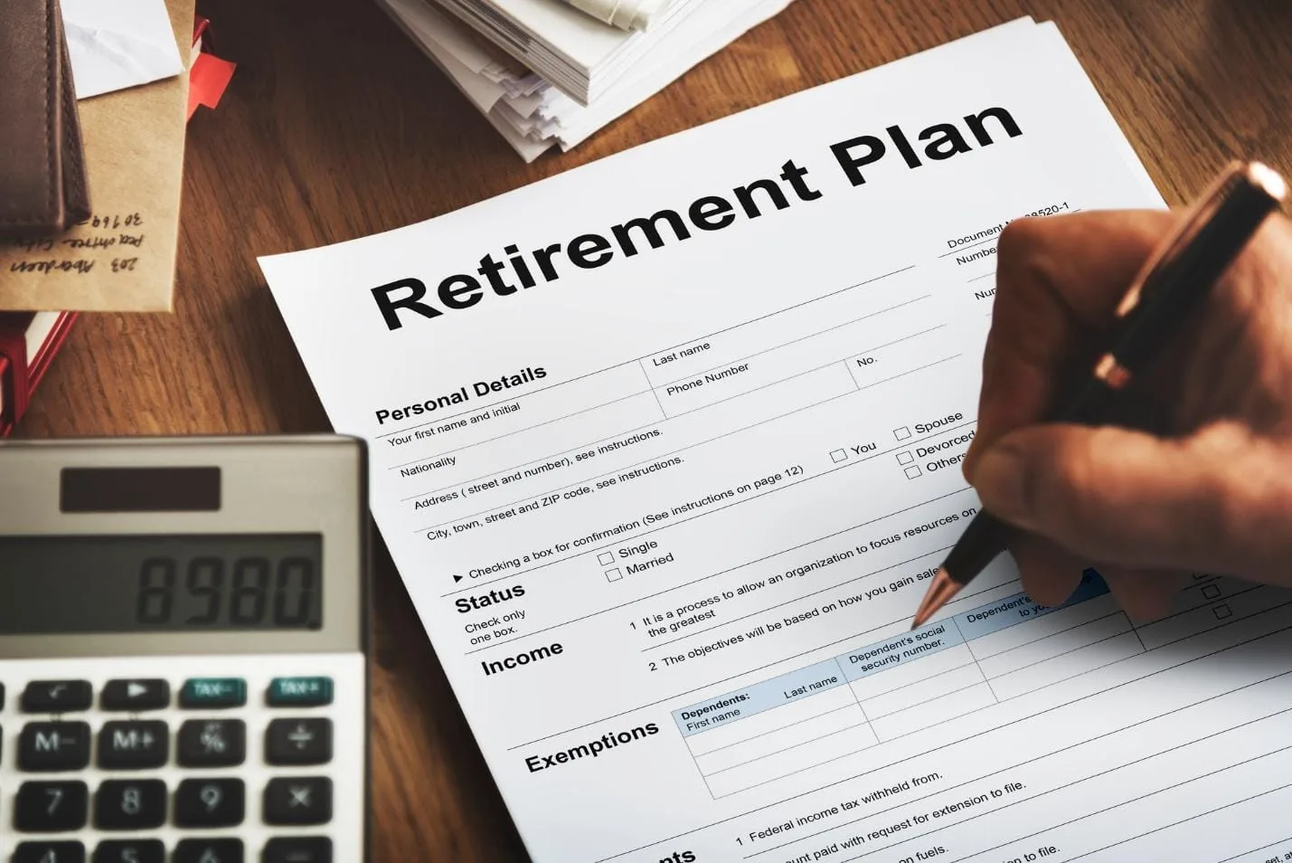 A calculator sits on a desk beside a paper that is titled Retirement Plan and a hand holding a pen hovers over the paper.