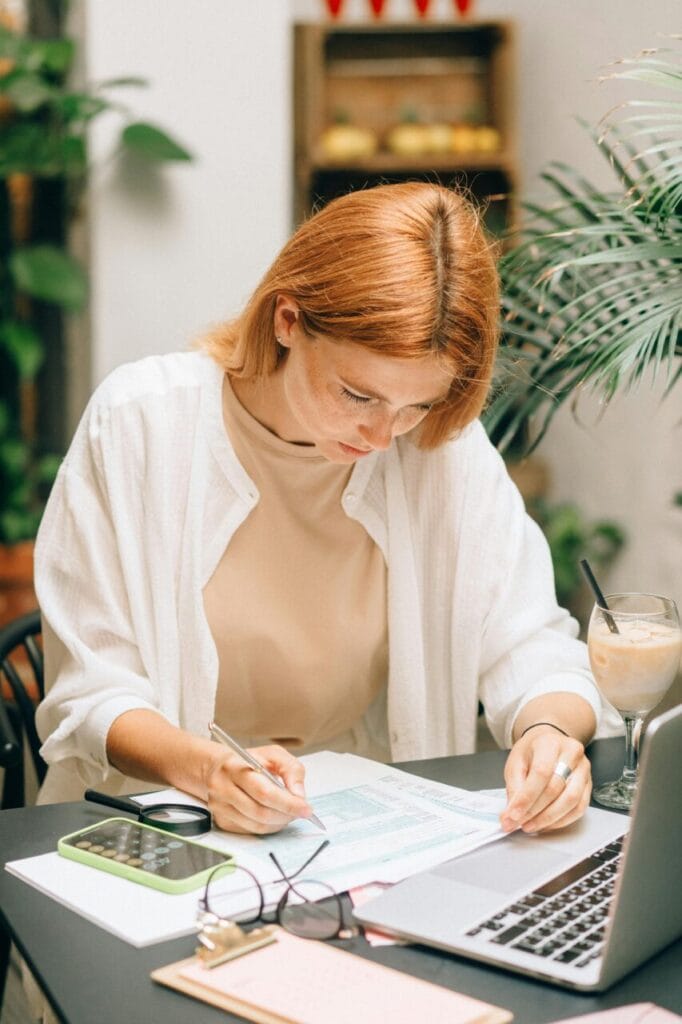 Focused woman with red hair working at a desk, reviewing documents with a laptop, phone, and drink nearby in a cozy setting with plants.