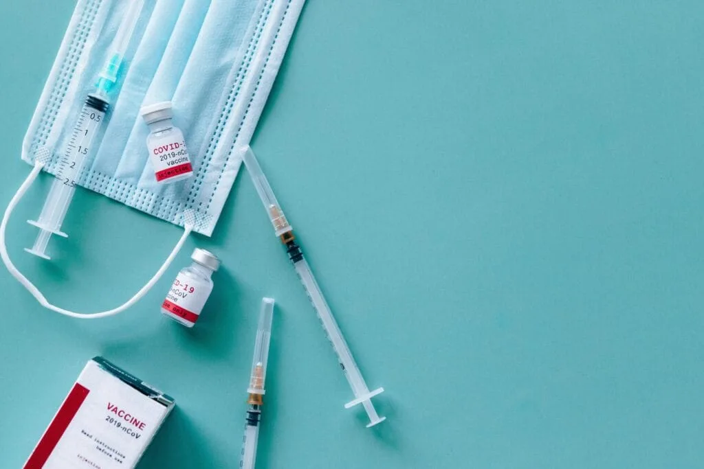 Medical items including syringe vaccine vials, a surgical mask, and a vaccine box arranged on a light blue surface.