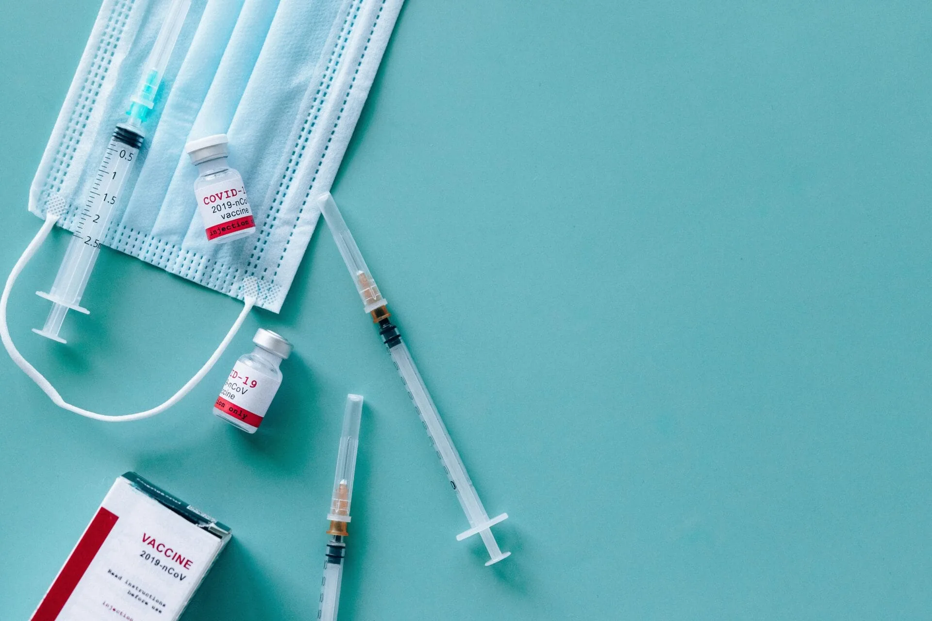 Medical items including syringe vaccine vials, a surgical mask, and a vaccine box arranged on a light blue surface.