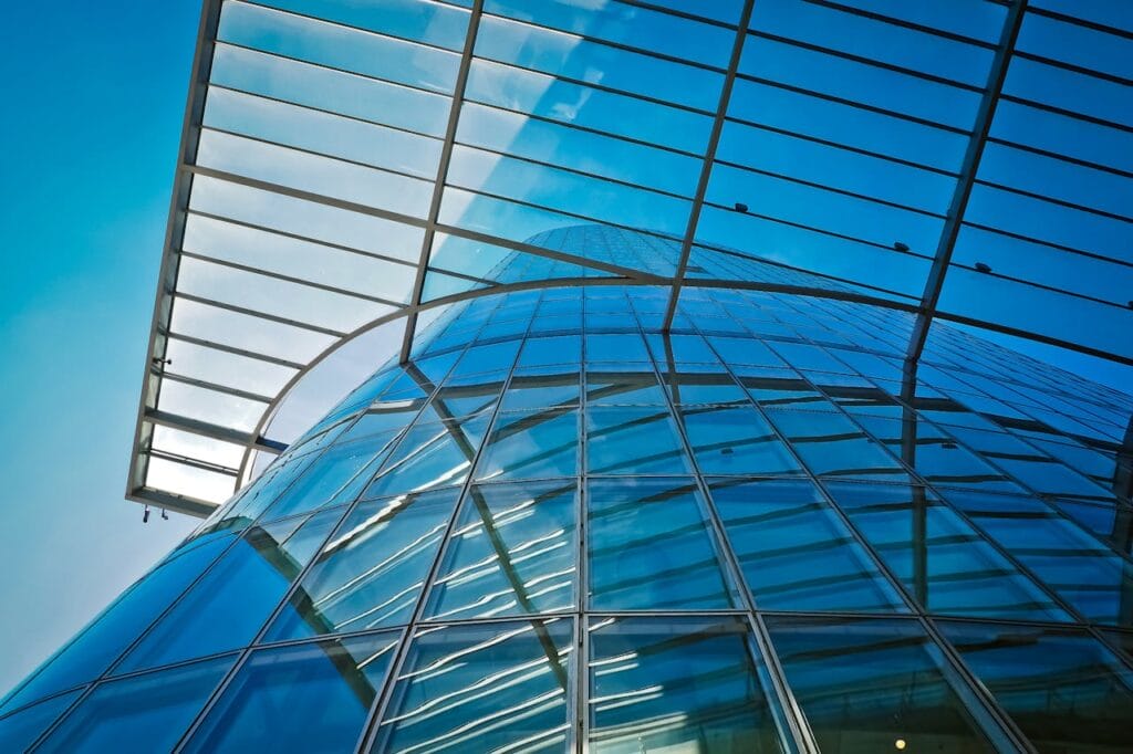 Low-angle view of a modern glass building with a curved facade and a metal framework under a bright blue sky.