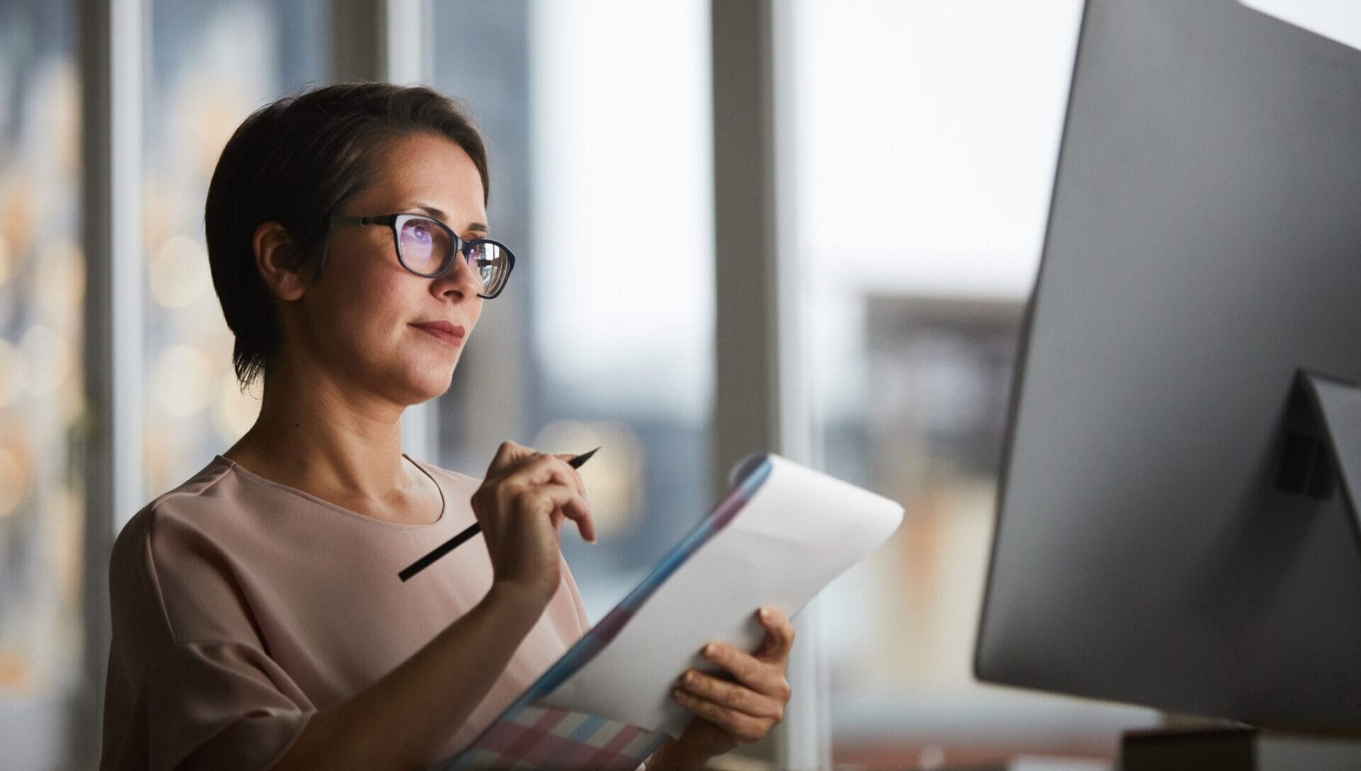 Confident businesswoman with document looking at computer screen and making working notes in office